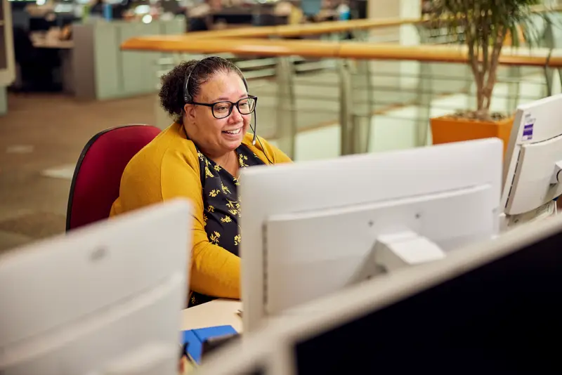 A woman in a yellow jumper sitting at a computer wearing a telephone headset