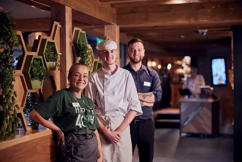 three employees standing against a wooden wall with plants in it. The lady at the front is in a green t-shirt. The Man in the middle is in chefs whites. The man at the back is in a blue shirt and black trousers