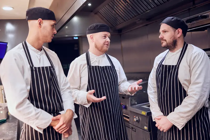 three employees standing against a wooden wall with plants in it. The lady at the front is in a green t-shirt. The Man in the middle is in chefs whites. The man at the back is in a blue shirt and black trousers