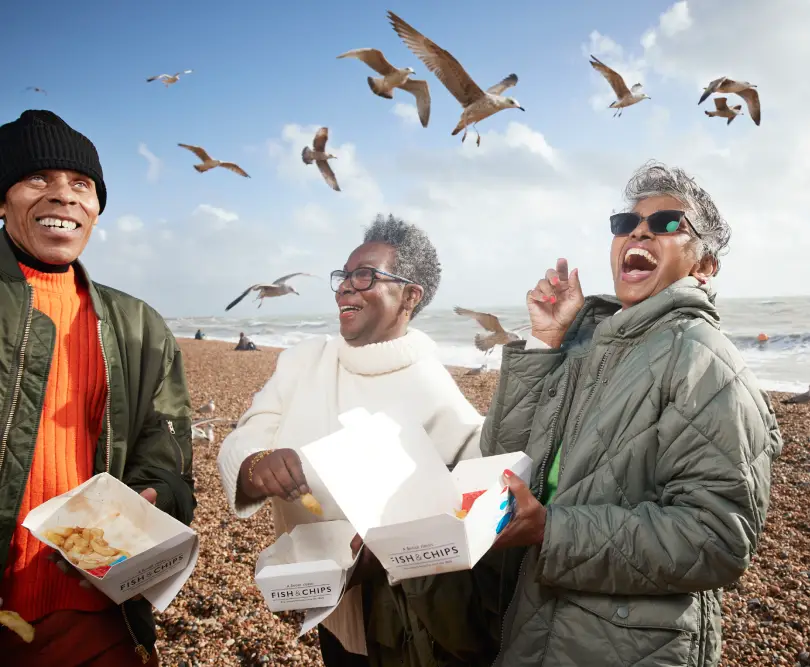 Three people on a pebbled beach eat fish and chips while seagulls fly overhead. The ocean is in the background under a partly cloudy sky.