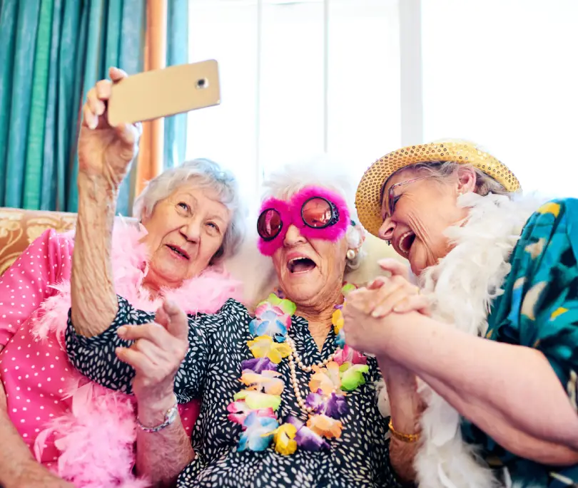 Three elderly women in festive attire, including feather boas and hats, take a selfie together while sitting on a couch.